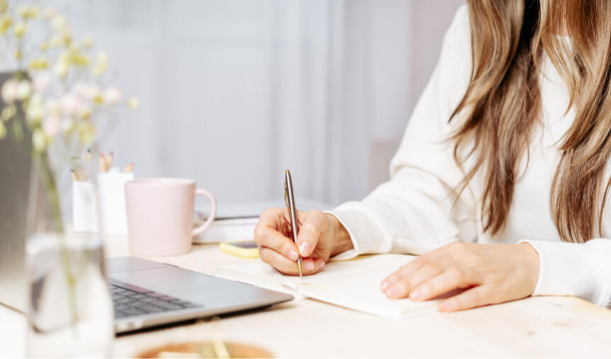 Let's write together. A female writer sits with a pen and paper in front of a computer.